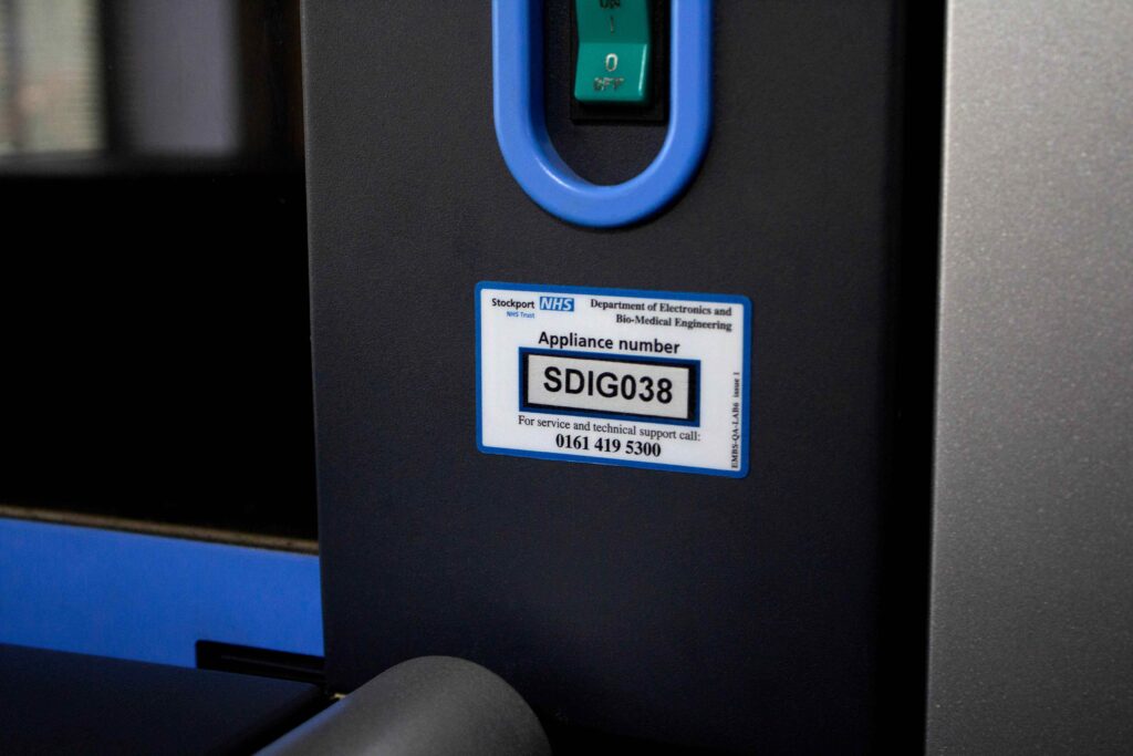 White and blue plastic asset label with black text stuck to electronic machine with clear window showing serial number